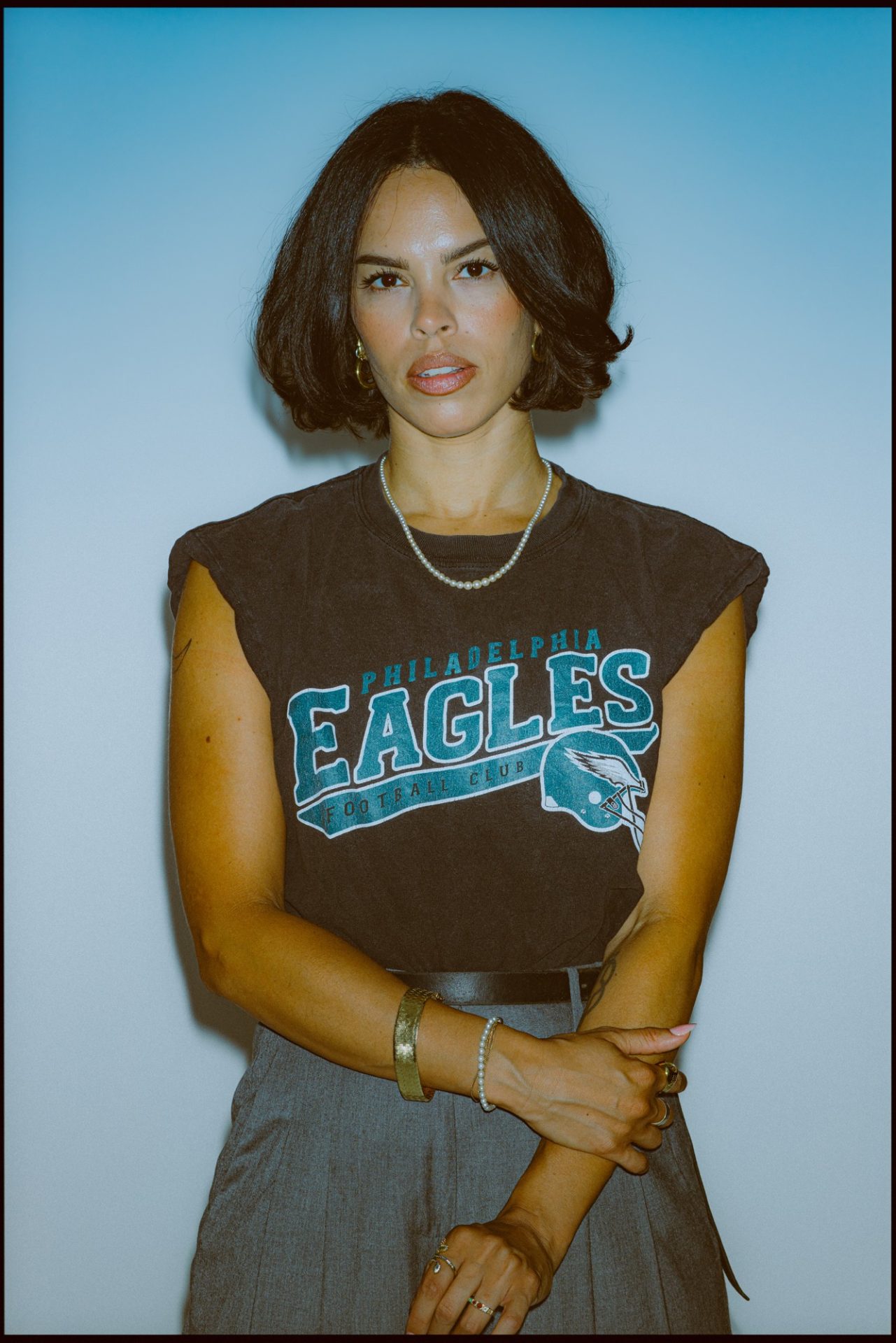 Zu sehen ist Sophie Yukiko vor einem blauen Hintergrund. Sie trägt ein braunes, ärmelloses Oberteil, auf dem ein Philadelphia Eagles-Schriftzug zu sehen ist. Dazu eine graue Hose. Mit ihrer rechten Hand greift sie sich an den linken Unterarm.