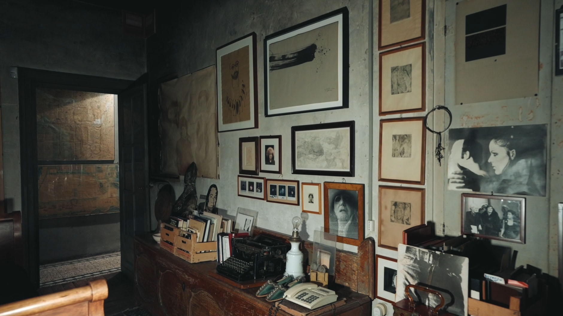 Blick auf eine Wand, an der gerahmte Malereien und Fotografien hängen. Darunter befindet sich eine Holzkommode, auf der eine Telefon, eine Schreibmaschine und Holzkisten mit Büchern stehen.